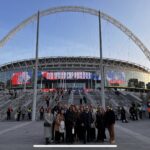 A group of people standing in front of Wembley Stadium, with the iconic arch visible in the background. The stadium is hosting a FIFA World Cup qualifier, as indicated by the signage, and security personnel and attendees are seen in the background near the entrance.