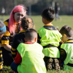 Sport England Image. Female coach sits with group of children bedside football pitch. Children wear black and yellow kits with high viz vests.