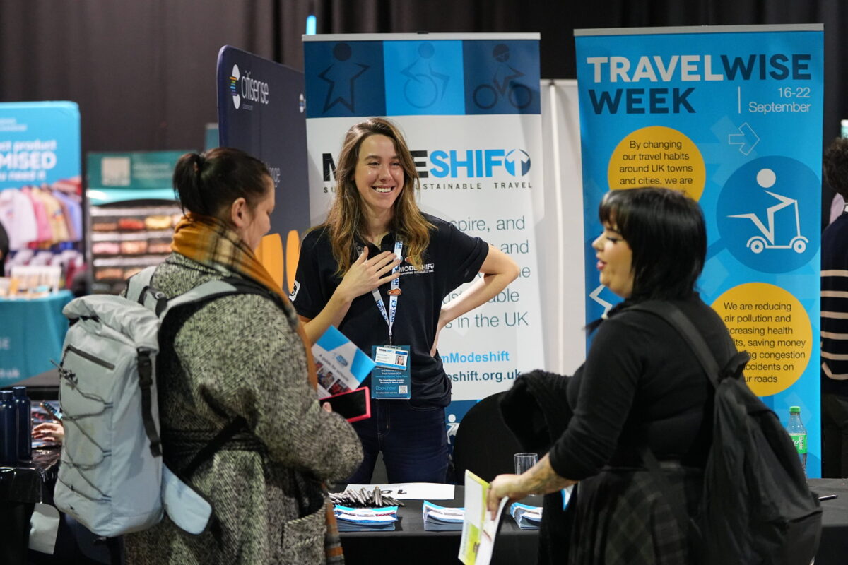 Three women engaged in conversation. The woman in the centre smiles, standing in front of a Modeshift branded display stand. A table with leaflets in front of her.