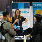 Three women engaged in conversation. The woman in the centre smiles, standing in front of a Modeshift branded display stand. A table with leaflets in front of her.