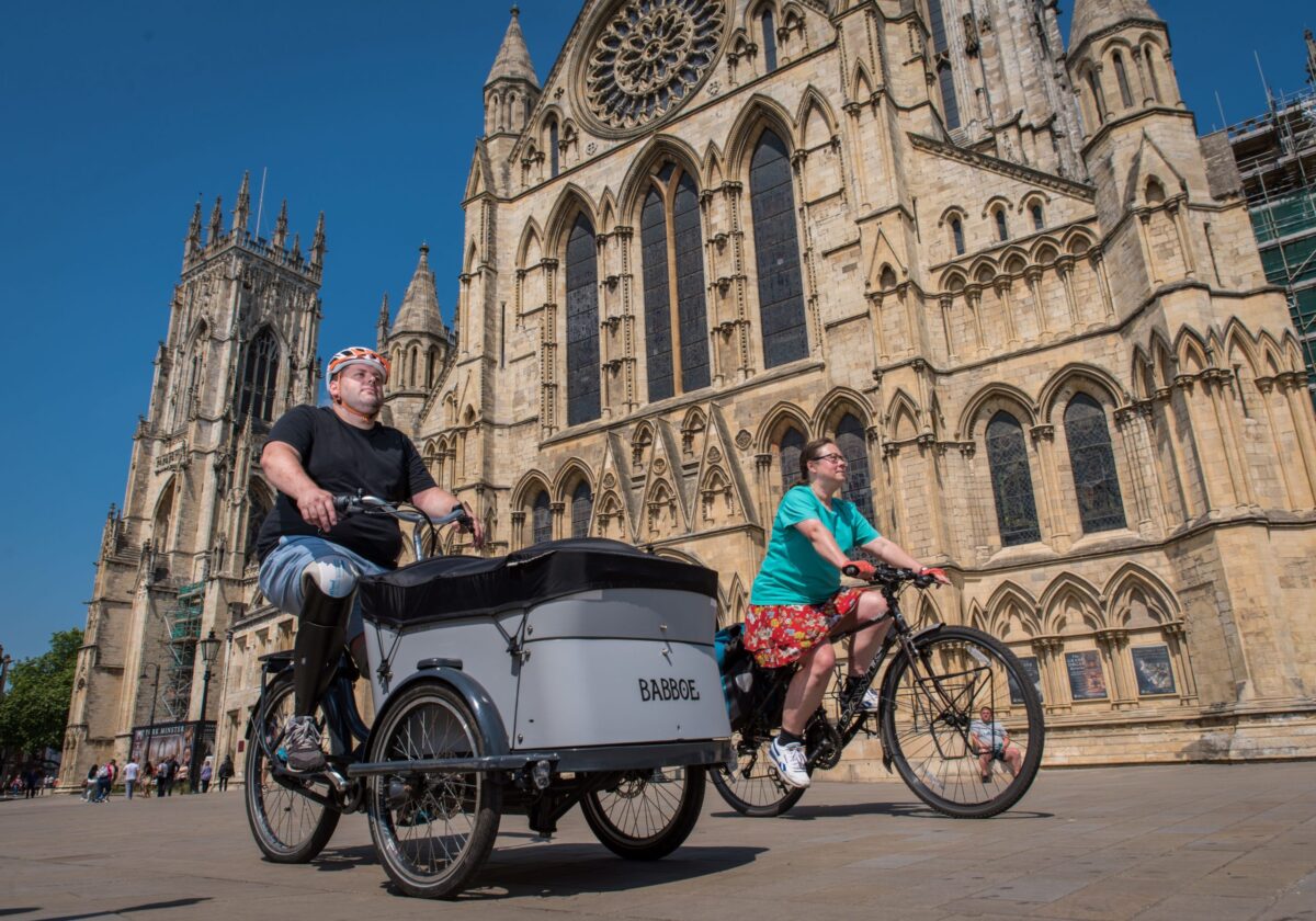 Image credit: Active Travel England. Two people cycle past York Minster. Person in the bottom left is peddling a Cargo Bike.