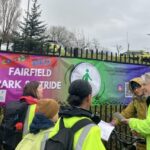 A group of people gather by a railing banner that reads: Fairfields Park and Stride. They wear high vis yellow vests.