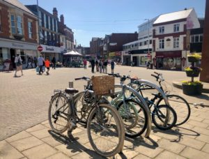 Four bikes parked in bike parking station in Kettering Town Centre, high street in background with shops and people walking.
