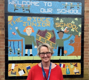 Image shows Beverly Furbur from The Ridge Primary school smiling infront of a sign wearing a red top