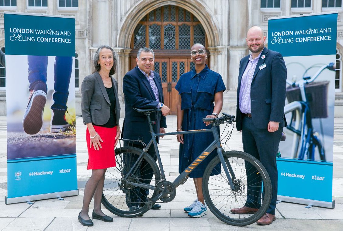 Four top speakers from the 2022 edition of the London Walking and Cycling Conference, standing outside the venue holding a modern road bike, standing next to two promotional roll-up banners