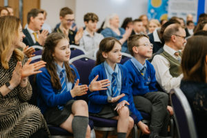 Group people seated in rows. Closest to the camera are three school children. They are wearing blue school jumpers with grey trousers and skirts/long socks. 