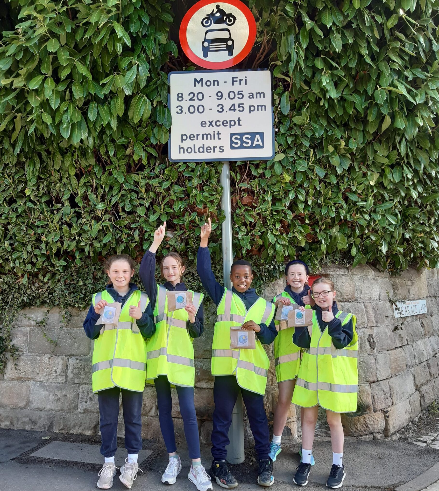 Five school children wearing green uniform and yellow high vis vests stand by a 'School Streets' sign that outlines times the road may be used by motorists. They are smiling and pointing up at the sign.