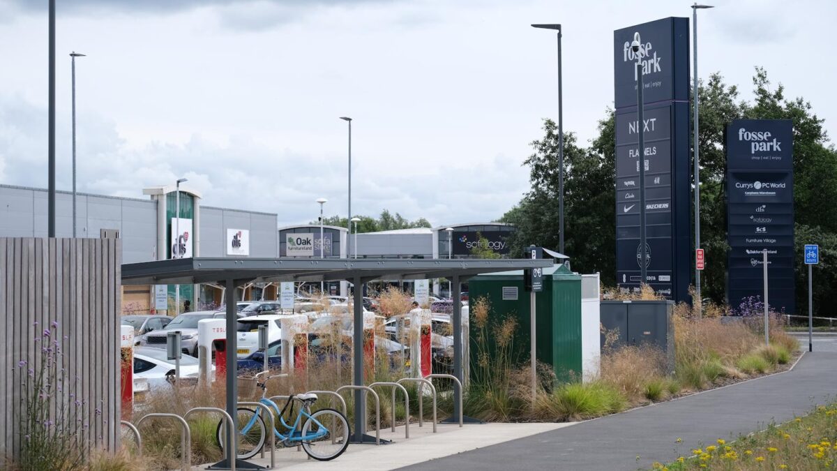 Fosse Park, a bicycle is parked in a covered shelter. In the back ground a shopping centre with large units can be seen, a car park in front of them with cars parked. A sign to the right reads: Fosse Park.