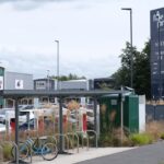 Fosse Park, a bicycle is parked in a covered shelter. In the back ground a shopping centre with large units can be seen, a car park in front of them with cars parked. A sign to the right reads: Fosse Park.