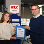 A woman and a man smiling and holding a framed Modeshift STARS Accreditation certificate. Behind them is a vintage-style petrol pump with "RIX PREMIUM" signage, and a modern office setting is visible in the background.