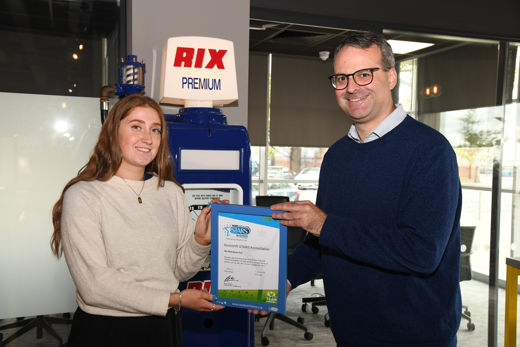 A woman and a man smiling and holding a framed Modeshift STARS Accreditation certificate. Behind them is a vintage-style petrol pump with "RIX PREMIUM" signage, and a modern office setting is visible in the background.