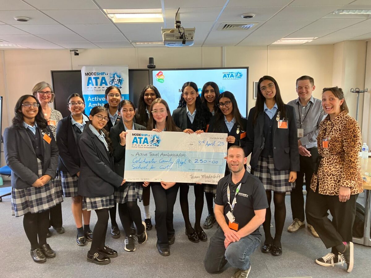 A group of students from Colchester County High School for Girls, along with school staff and Modeshift representatives, smile as they hold a large £250 cheque for their Active Travel Ambassador campaign. A Modeshift ATA banner is visible in the background.