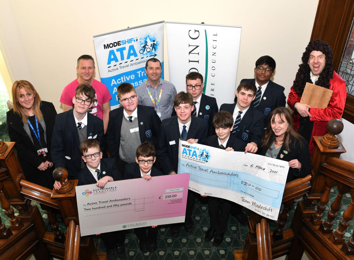 A school group of boys in uniform pose on a staircase alongside teachers and supporters, holding two large cheques for £250 each. The scene is part of an Active Travel Ambassador event in East Riding, with Modeshift ATA banners behind them.