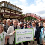 A group of people stand on a railway platform, smiling and holding a sign that reads: We're proud to support Community Rail Week