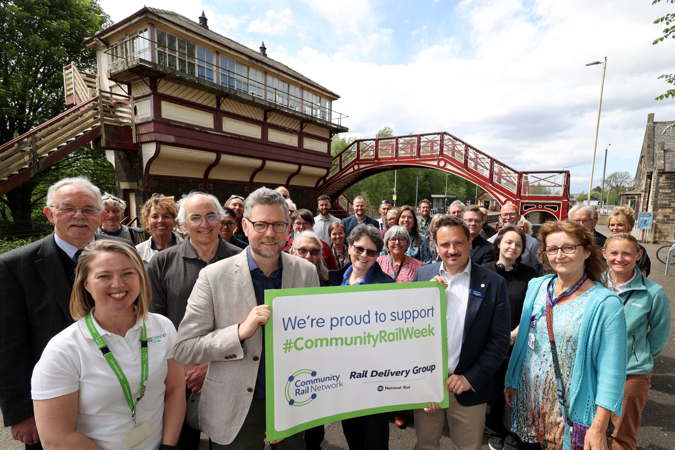 A group of people stand on a railway platform, smiling and holding a sign that reads: We're proud to support Community Rail Week