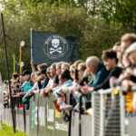 A crowd of football supporters lined up along the sideline during a daytime match. A black flag featuring the Real Bedford FC skull and crossbones logo waves prominently above the fans. How football clubs reduce matchday traffic.