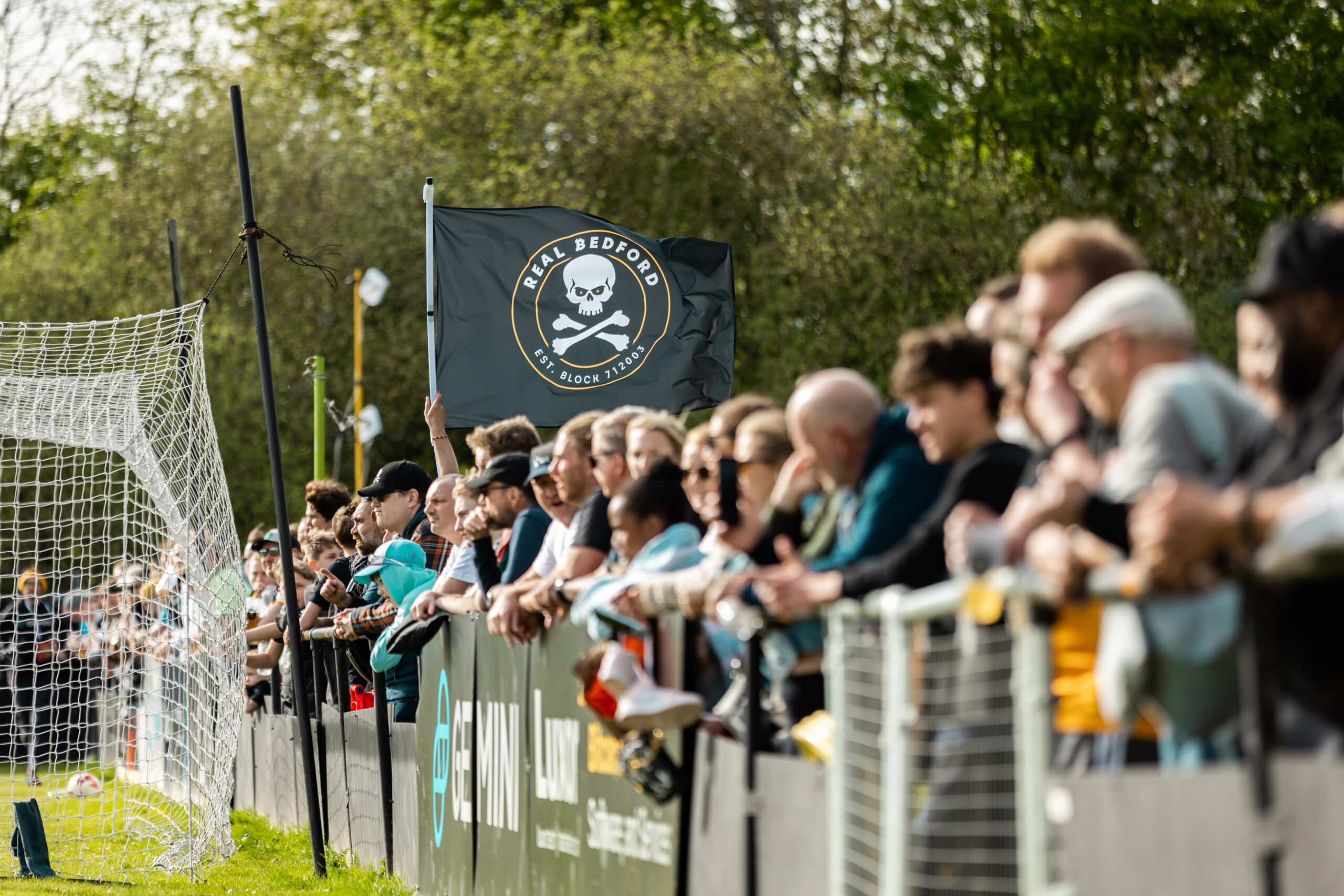 A crowd of football supporters lined up along the sideline during a daytime match. A black flag featuring the Real Bedford FC skull and crossbones logo waves prominently above the fans. How football clubs reduce matchday traffic.