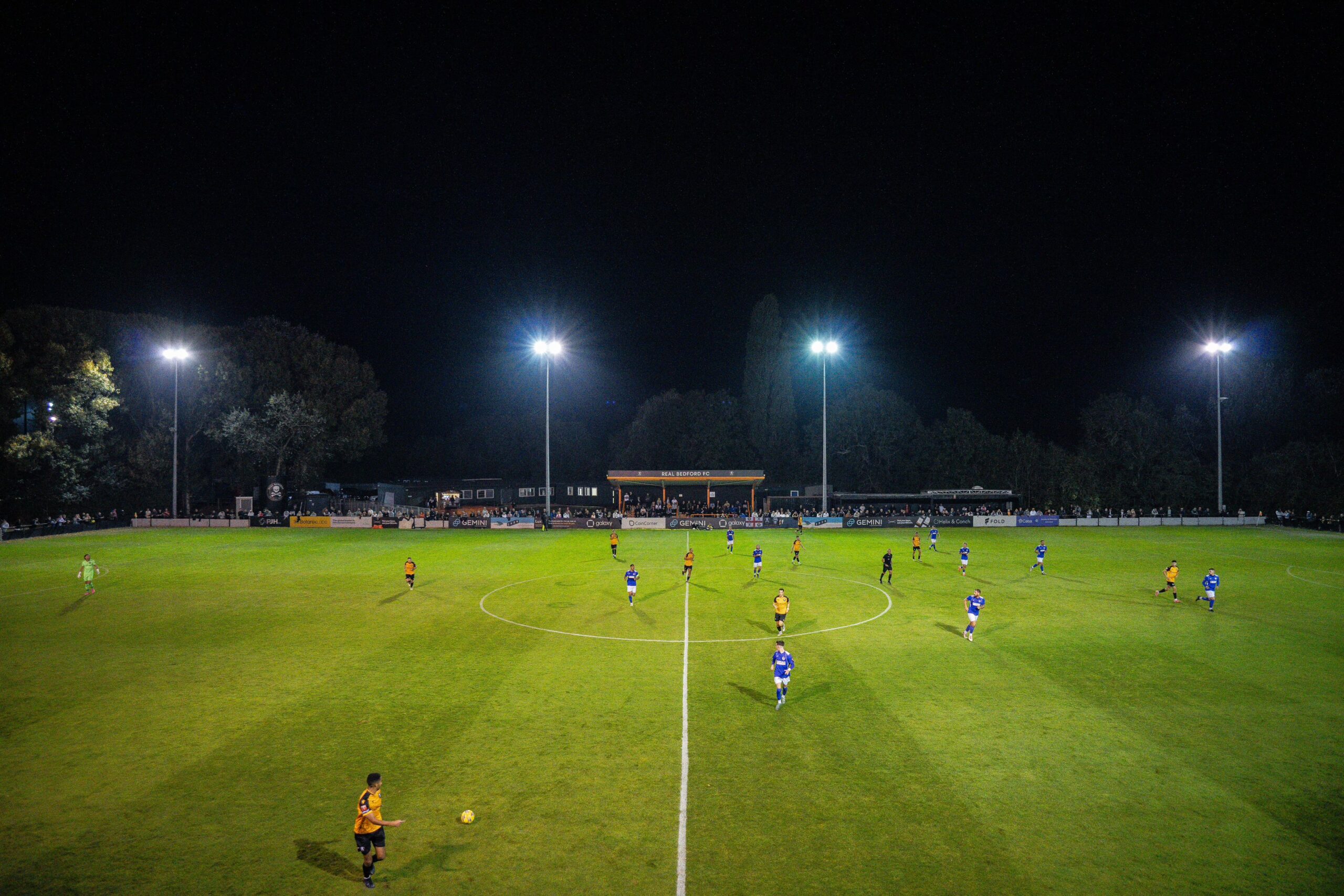 A night-time football match in progress under floodlights at a packed stadium. Players in orange and blue kits are spread across a lush green pitch, with fans filling the stands in the background.