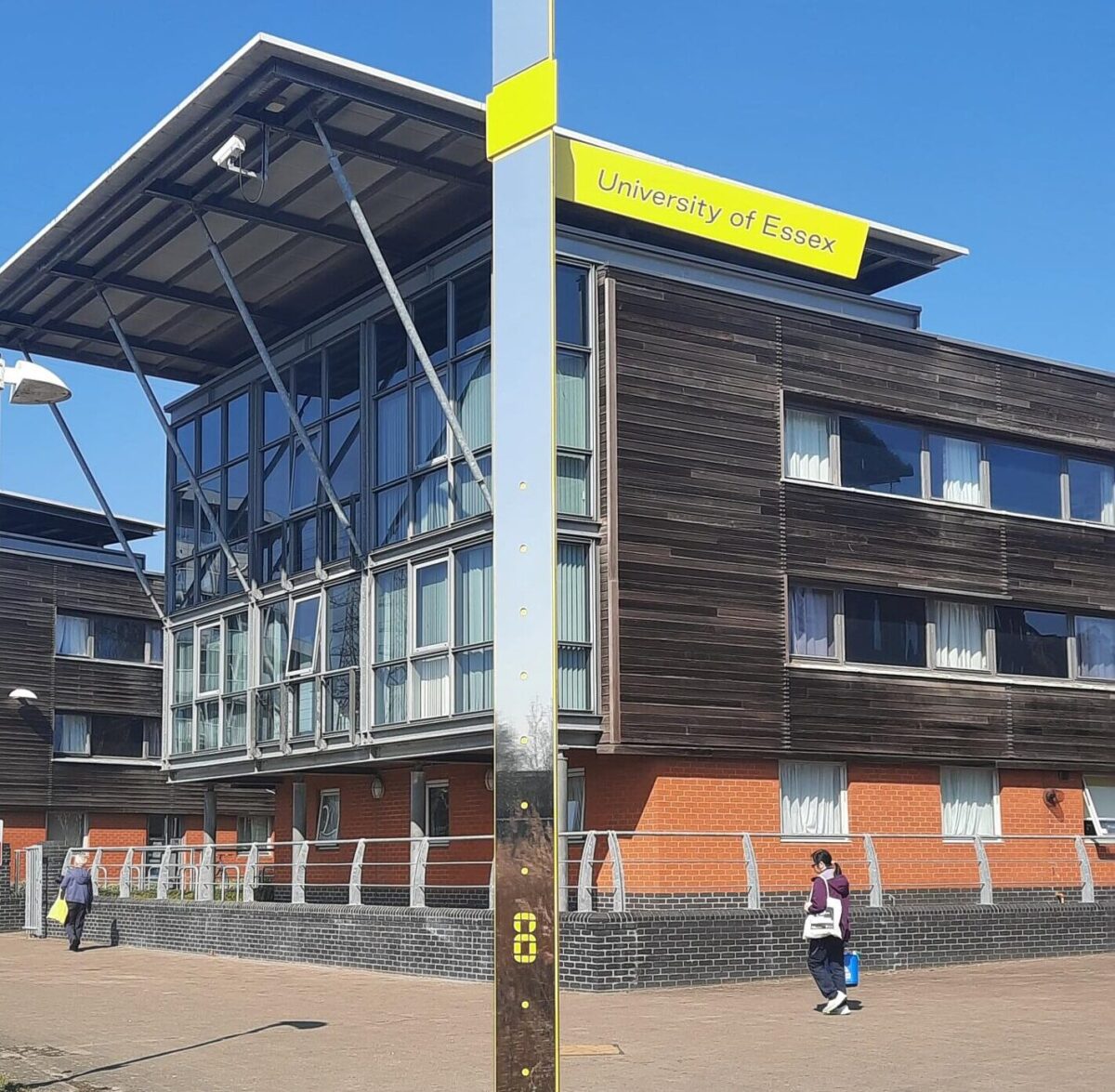 Large building - The University of Essex. A sign post reads University of Essex in front of the building. People ca be seen walking by the building. beneath a bright blue sky.