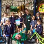 A group of school children and their teacher pose for the camera with their bikes and scooters. A school crossing sign is behind them.