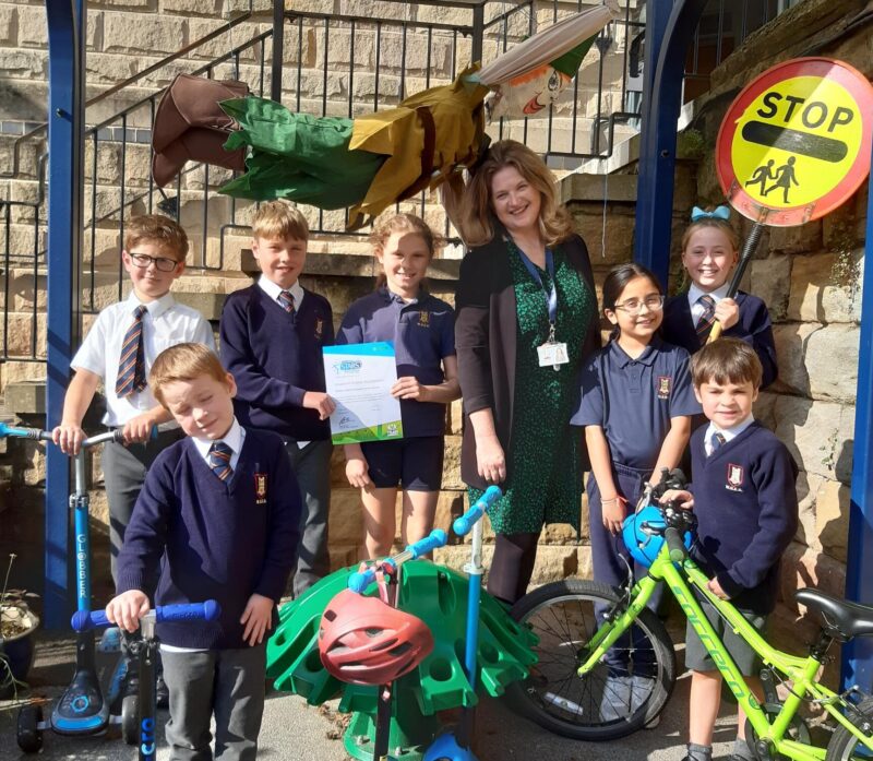 A group of school children and their teacher pose for the camera with their bikes and scooters. A school crossing sign is behind them.