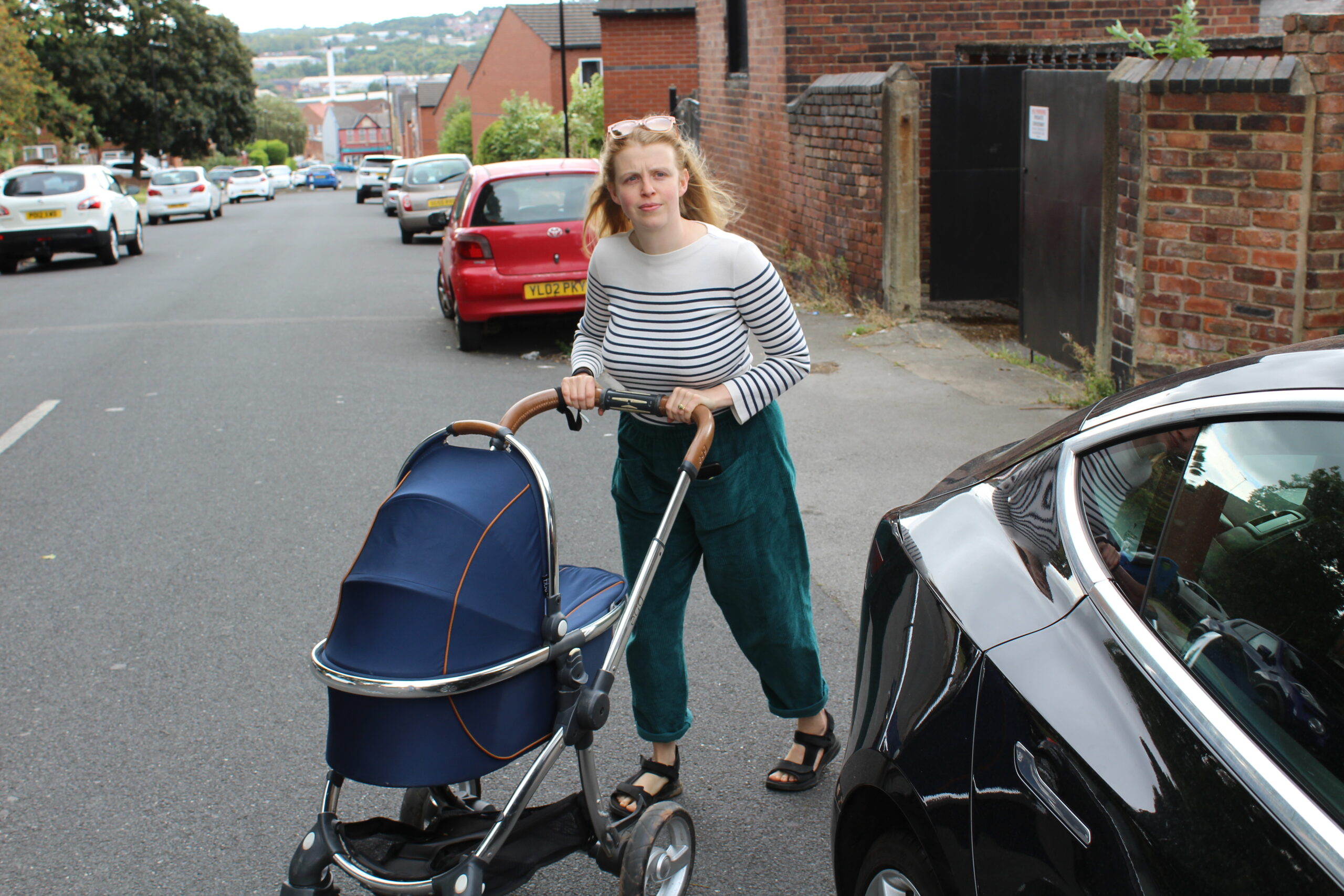 A woman pushing a pram is forced to walk in the road due to a car parked on the pavement. She looks concerned as she navigates between parked vehicles on a residential street. Image credit: Living Streets