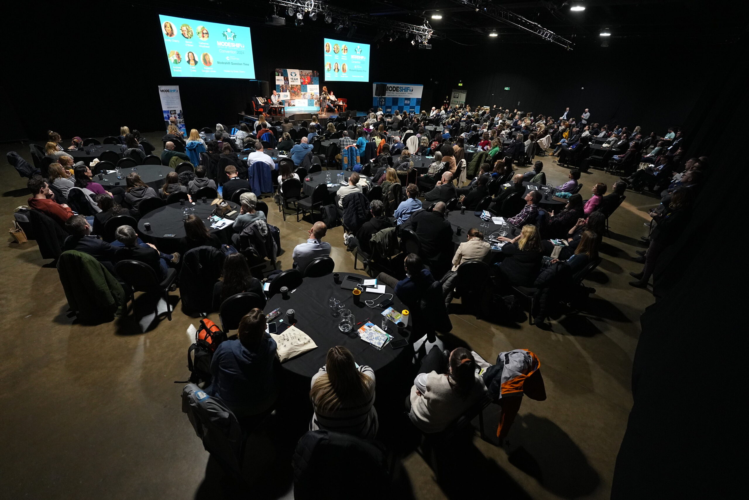 A large meeting room with people seated around round tables. A stage is the focus of the audience attention with two large screens displaying Modeshift branded presentations.