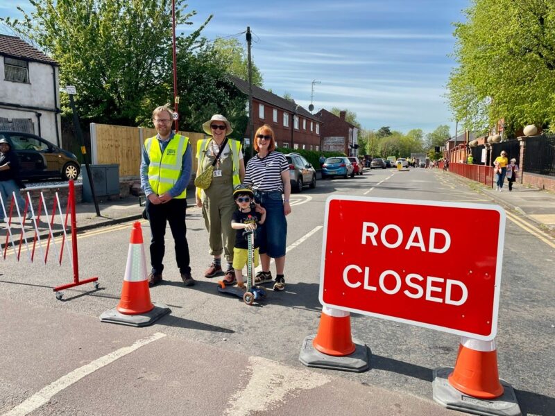 Man and two woman and child on a scooter wearing yellow high vis jackets at a road closed sign at the end of a street 