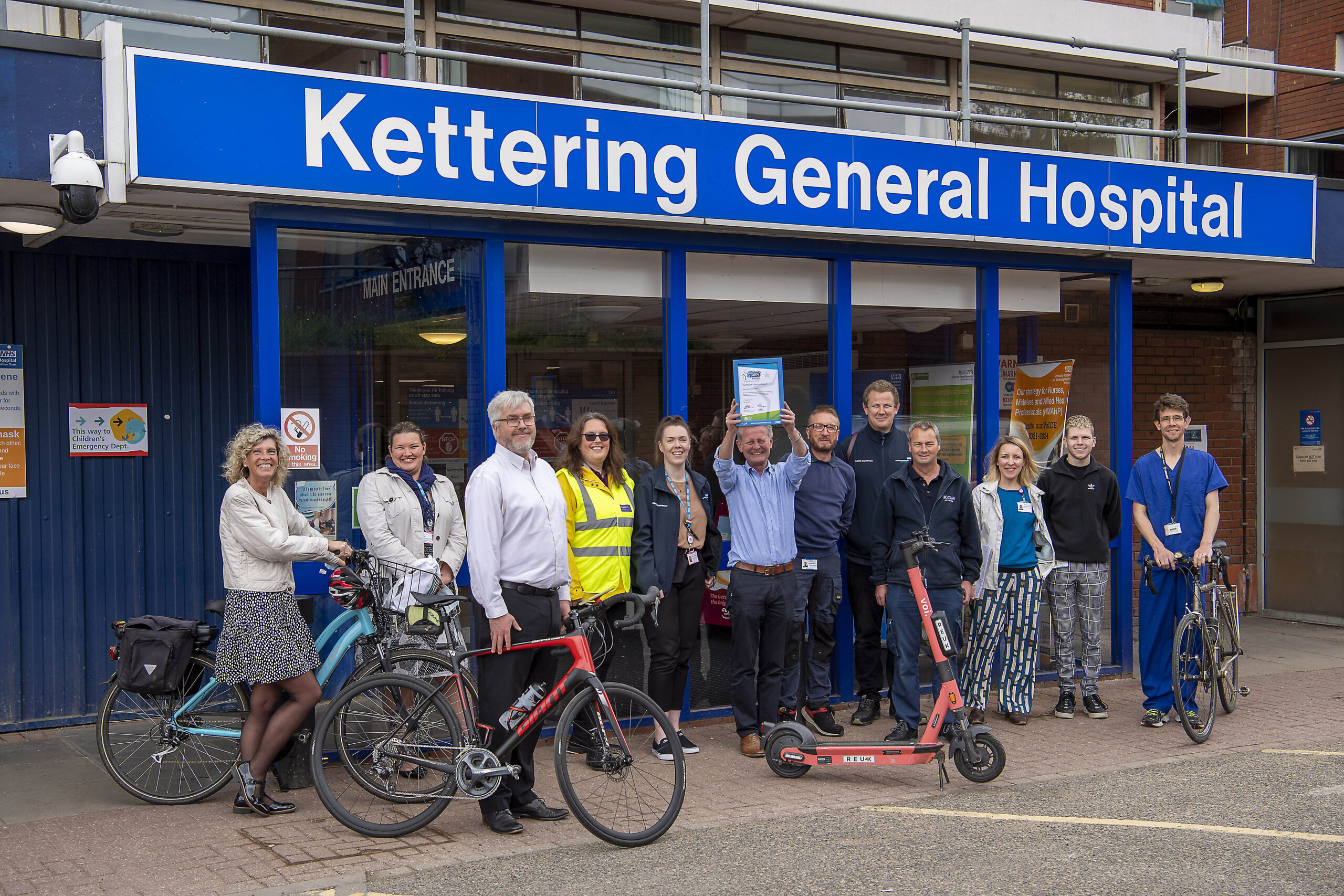 A large group of people gather by the entrance to Kettering General Hospital. Some have bicycles and a man in the centre holds up a Modeshift STARS Certificate.