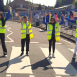 five children stood on a crossing smiling with arms in the air wearing yellow high vis jackets