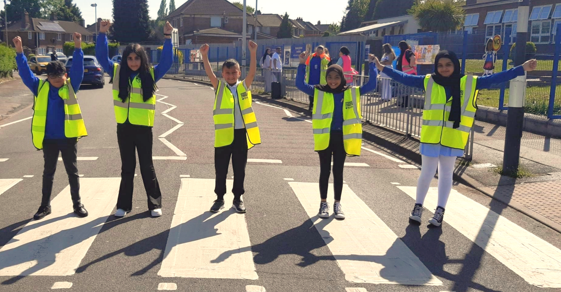 five children stood on a crossing smiling with arms in the air wearing yellow high vis jackets
