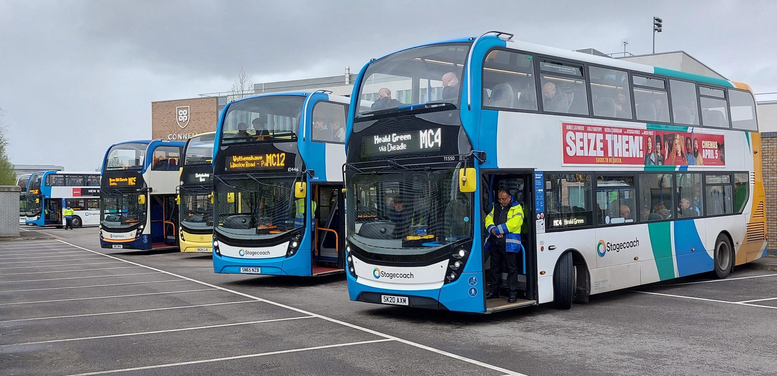 Multiple Stagecoach double-decker buses are lined up outside Connell Co-op College, ready for service. The destination signs display various matchday routes such as MC4 to Heald Green and MC12 to Wythenshawe. A driver in a high-visibility jacket stands at the door of one bus as passengers are visible seated inside.