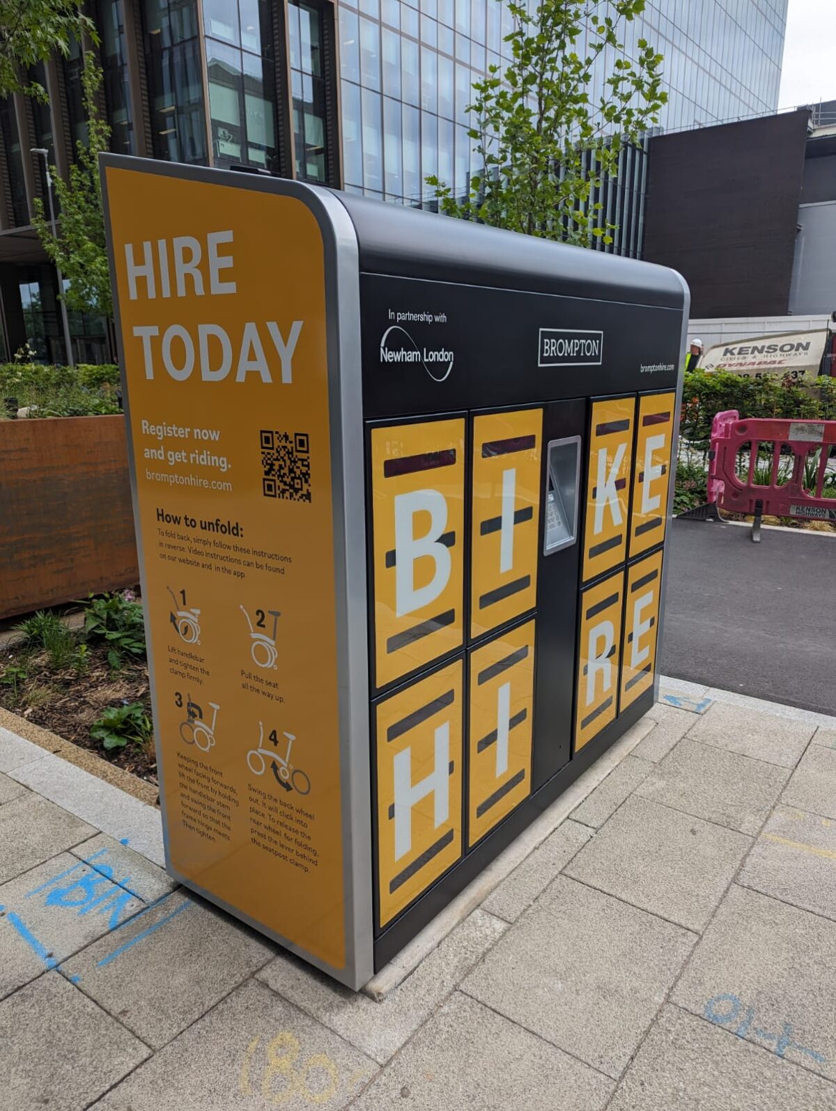 A Brompton Bike Hire dock located on a city pavement, branded in bright yellow and black with large letters spelling “BIKE HIRE.” The dock features individual lockers for folding bikes and a QR code to register. Instructions for unfolding a bike are displayed on the side. The dock is installed in partnership with Newham London and is situated near office buildings and urban greenery.