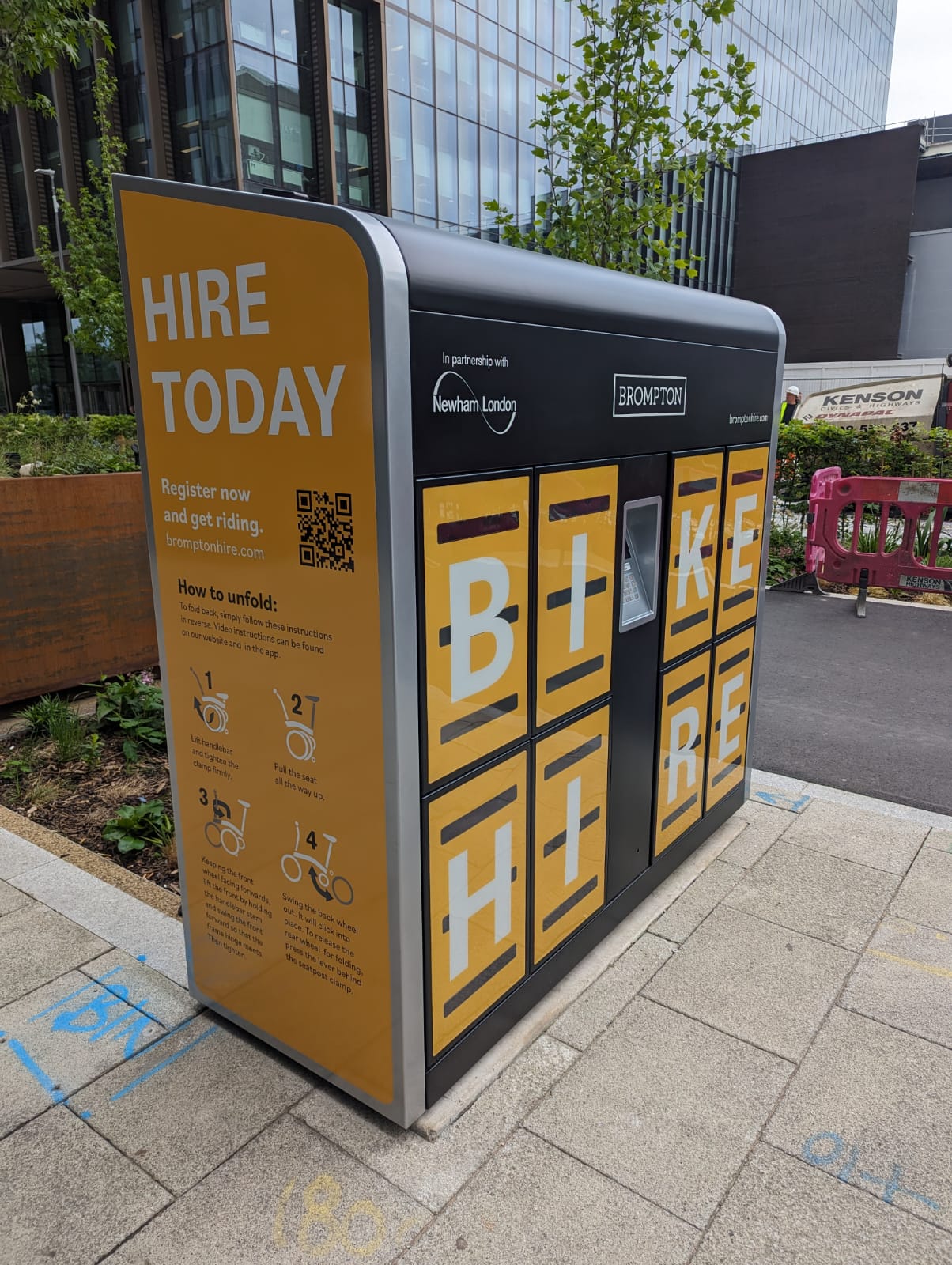 A Brompton Bike Hire dock located on a city pavement, branded in bright yellow and black with large letters spelling “BIKE HIRE.” The dock features individual lockers for folding bikes and a QR code to register. Instructions for unfolding a bike are displayed on the side. The dock is installed in partnership with Newham London and is situated near office buildings and urban greenery.