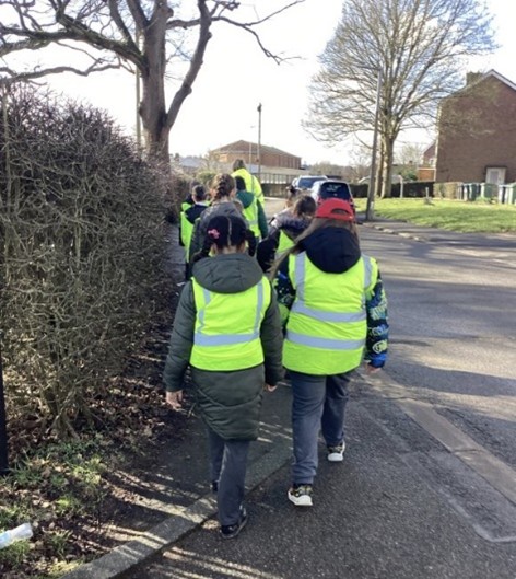 Picture shows children walking wearing high vis jackets walking at the side of a road with bushes and tree to the left and road on their right.