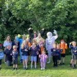 A cheerful group of children and adults dressed in space-themed costumes pose for a photo outdoors in front of leafy trees. Some children wear school uniforms, while others sport helmets, scooters, and bright accessories. One adult is dressed as an astronaut, and others wear alien or star costumes, celebrating a themed active travel event.
