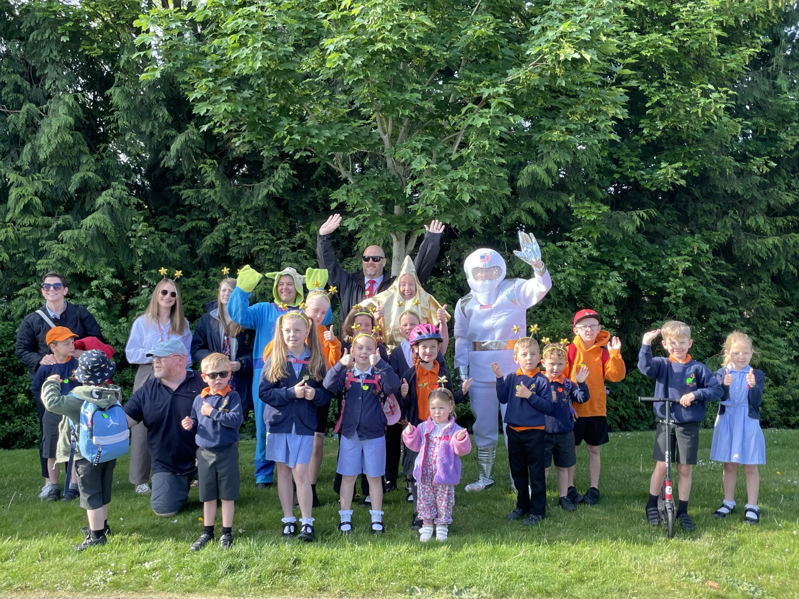 A cheerful group of children and adults dressed in space-themed costumes pose for a photo outdoors in front of leafy trees. Some children wear school uniforms, while others sport helmets, scooters, and bright accessories. One adult is dressed as an astronaut, and others wear alien or star costumes, celebrating a themed active travel event.