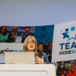 A woman speaks at a podium during the Modeshift Convention, with a large TEAM Modeshift banner behind her displaying photos of active travel and award moments.