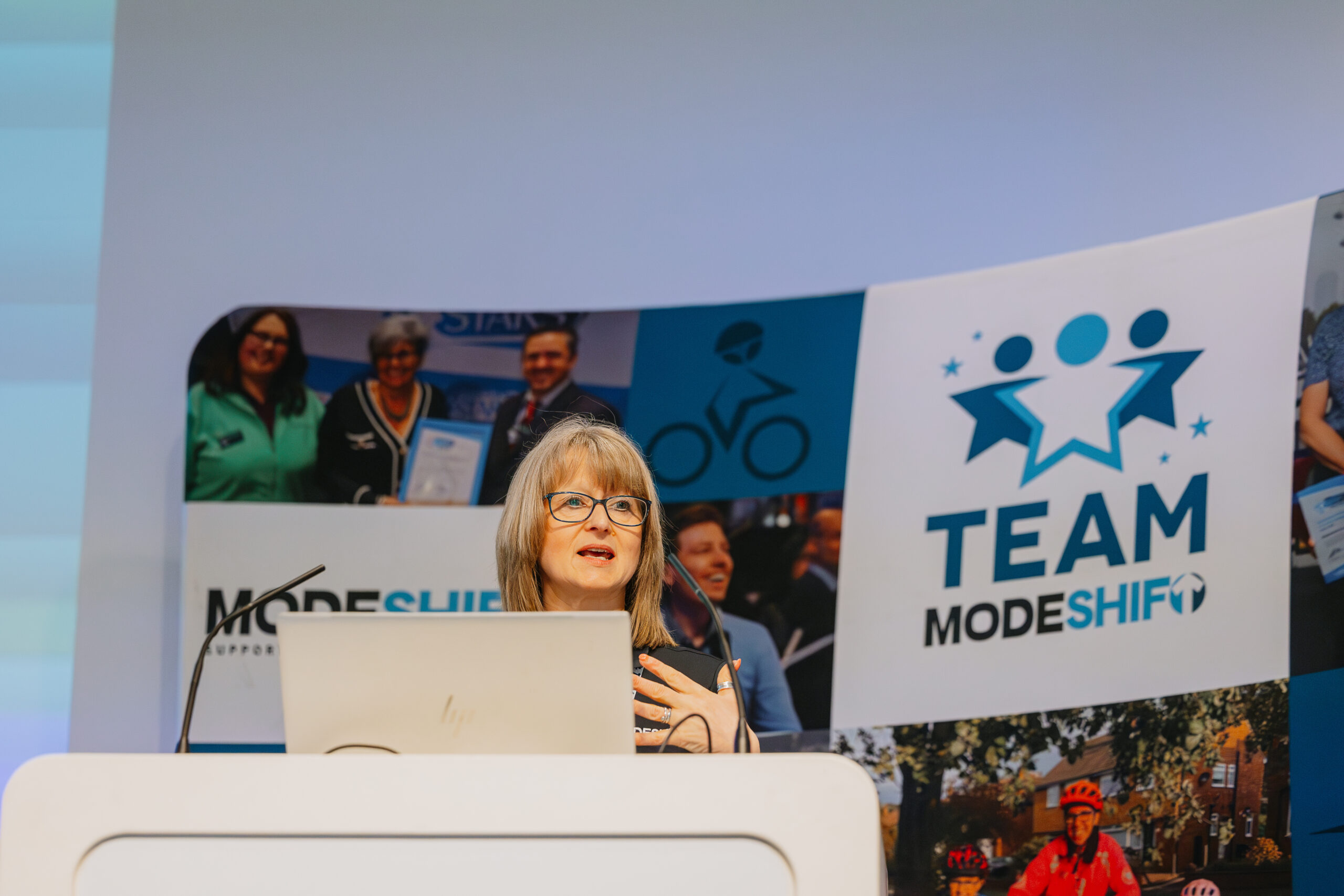 A woman speaks at a podium during the Modeshift Convention, with a large TEAM Modeshift banner behind her displaying photos of active travel and award moments.