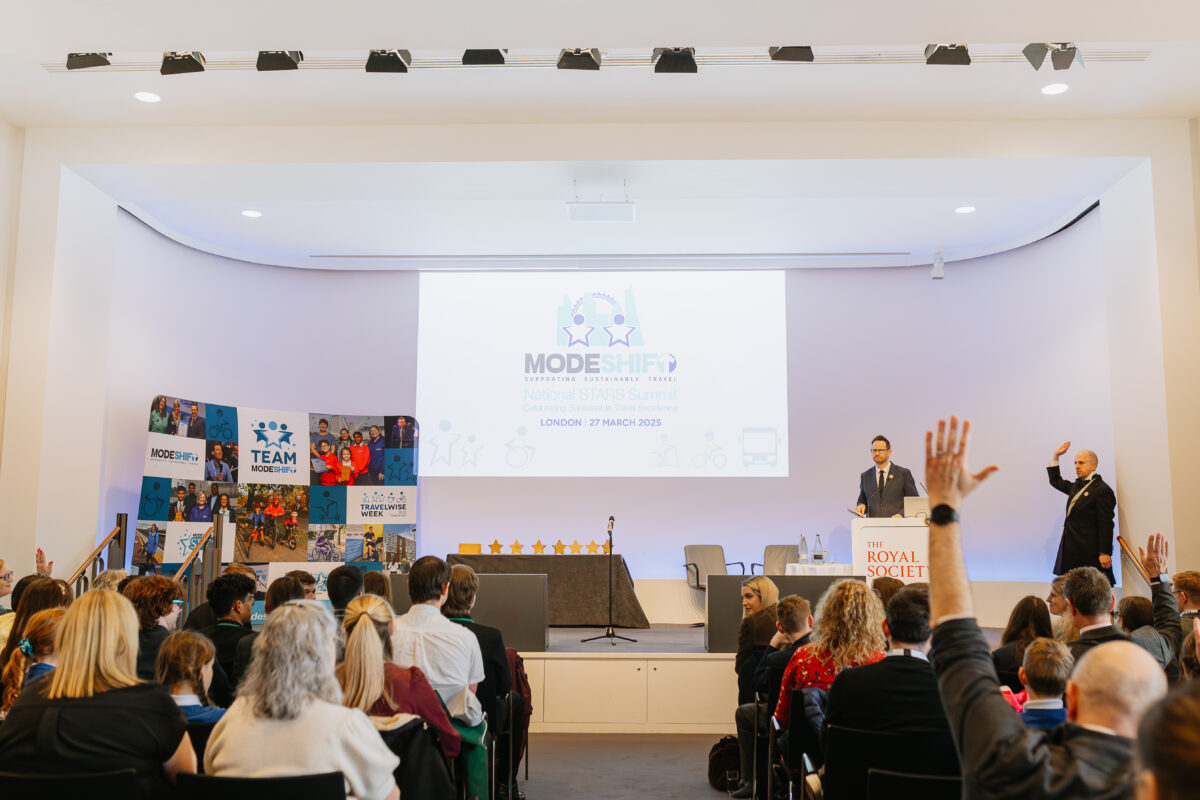 A speaker stands at a podium labeled "The Royal Society" during the 2025 Modeshift National STARS Summit in London, with a large presentation screen behind him displaying the event title and date. Attendees, including children and adults, sit facing the stage, with several raising their hands. A second presenter stands to the side, also engaging with the audience. A colorful display featuring Modeshift branding and photos is visible on the left side of the stage.