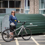 A woman stands confidently beside her bicycle in front of a green bike storage pod outside a brick building. She is wearing sunglasses, a blue top, a staff lanyard, and holding a bike helmet.