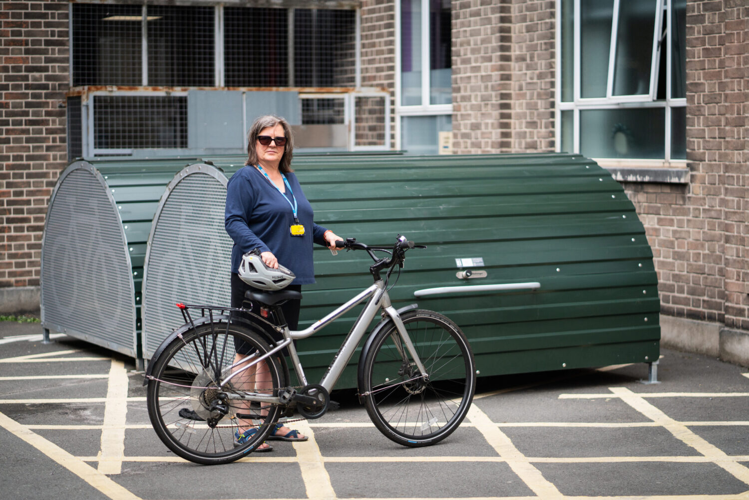 A woman stands confidently beside her bicycle in front of a green bike storage pod outside a brick building. She is wearing sunglasses, a blue top, a staff lanyard, and holding a bike helmet.