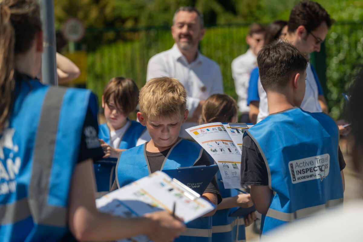 Children wearing blue vests labeled 'Active Travel Inspectors' reviewing clipboards outdoors during a group activity. How Active Travel Inspectors Support TfL's Walking and Wheeling Action Plan
