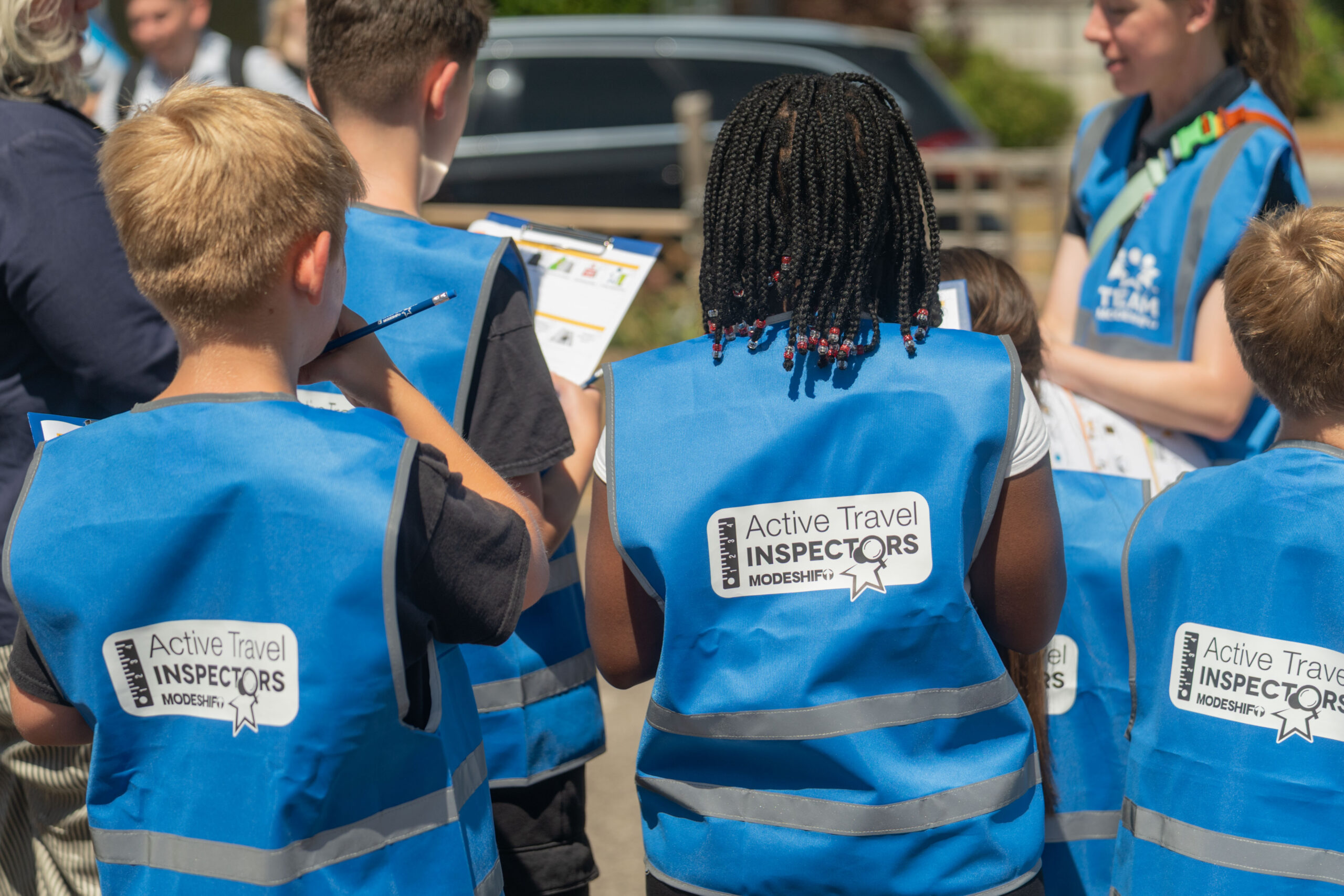 A group of children photographed from behind, wearing blue high viz vests with the Active Travel Inspectors logo on