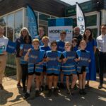 Group of children and adults wearing blue vests holding clipboards labeled 'Active Travel Inspectors' outside a building with banners promoting sustainable travel.