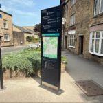 A pedestrian wayfinding totem stands on a pavement in Otley, featuring a map and local points of interest. It's located near a street corner with stone buildings, shops, and a directional signpost pointing to nearby attractions. A quiet road and greenery surround the area on a sunny day.