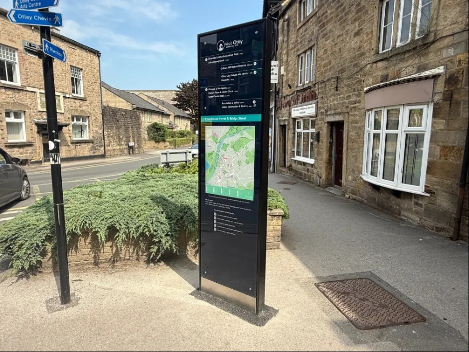 A pedestrian wayfinding totem stands on a pavement in Otley, featuring a map and local points of interest. It's located near a street corner with stone buildings, shops, and a directional signpost pointing to nearby attractions. A quiet road and greenery surround the area on a sunny day.