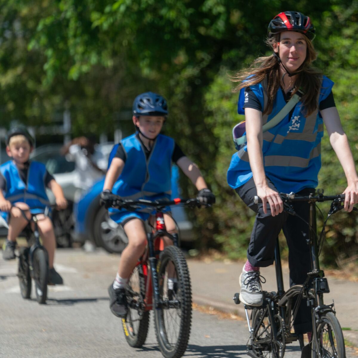 A woman rides a bike, wearing a cycle helmet and blue high viz top, a group of children wearing similar clothing cycle behind her.