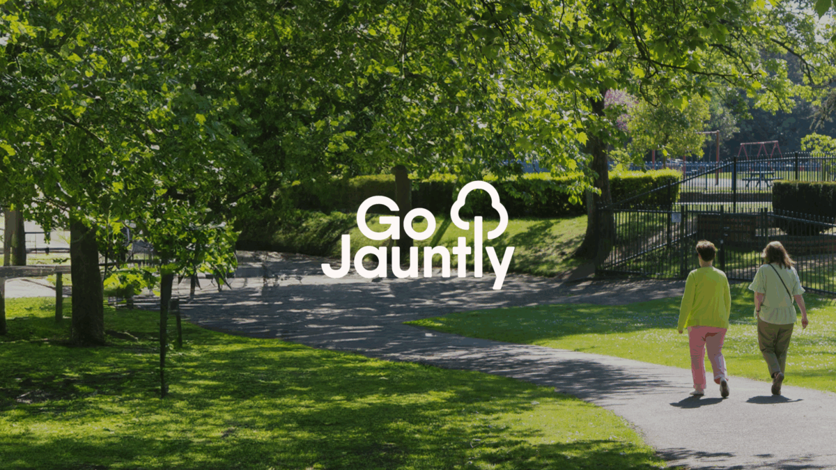 Two people walking on a shaded park path surrounded by lush green trees and grass on a sunny day. Go Jauntly logo in the centre