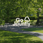 Two people walking on a shaded park path surrounded by lush green trees and grass on a sunny day. Go Jauntly logo in the centre